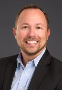 A person is smiling, wearing a suit and a blue shirt, against a gray background.