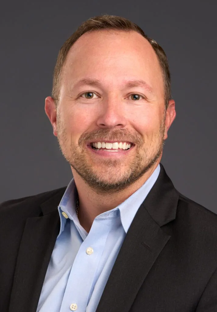 A person is smiling, wearing a suit and a blue shirt, against a gray background.