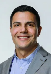A person smiling, dressed in a suit and blue shirt, wearing a beaded necklace.