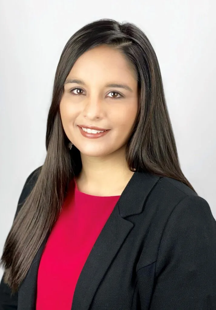 A person is dressed in a black blazer and red top, smiling against a plain backdrop.