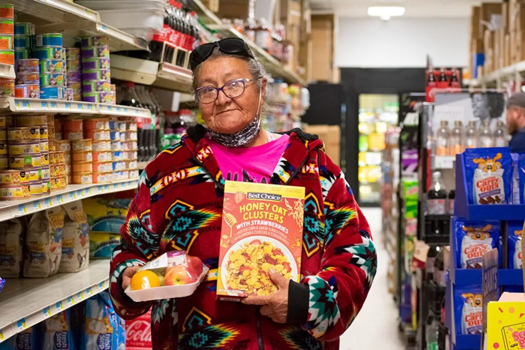 A person in a store holds a cereal box and a tray of apples, wearing a colorful patterned jacket.