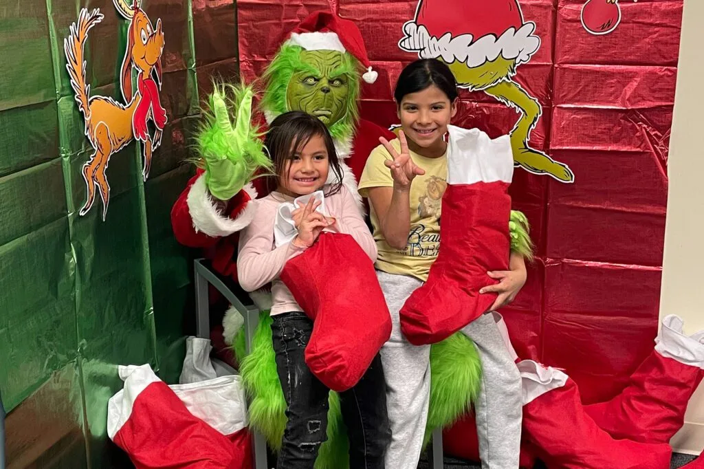 Two children pose with someone dressed as the Grinch, holding Christmas stockings.