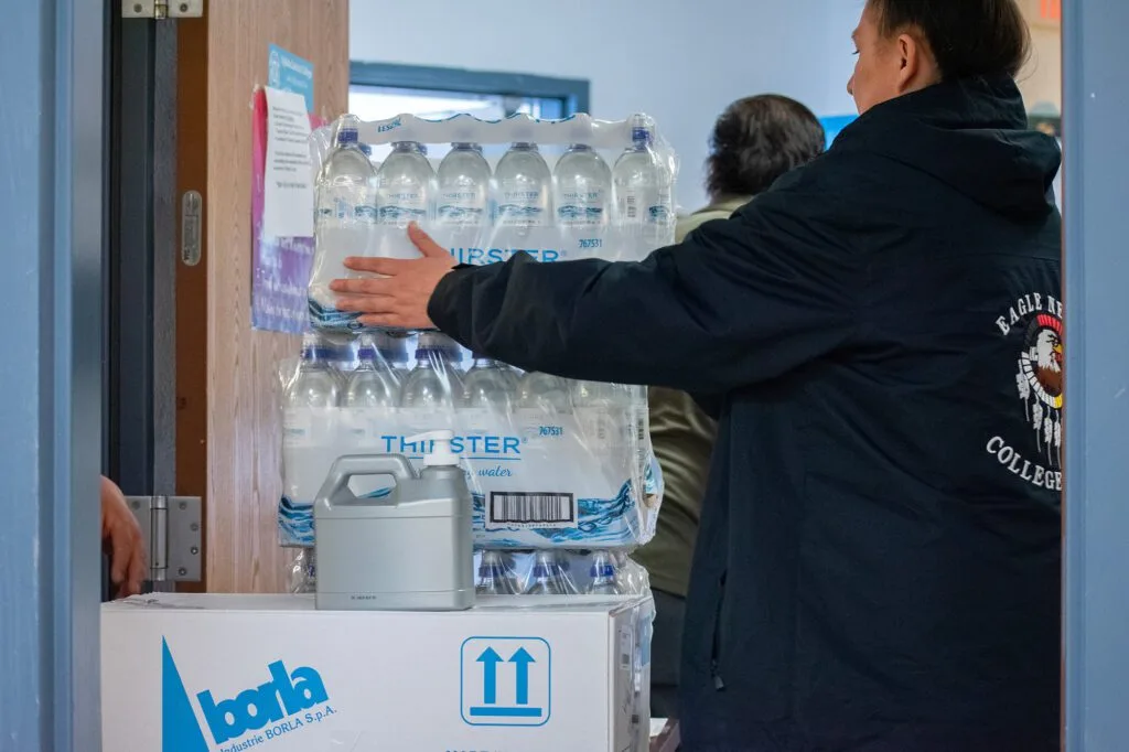 People are carrying a stack of bottled water through a doorway, possibly related to northern plains reservation aid.