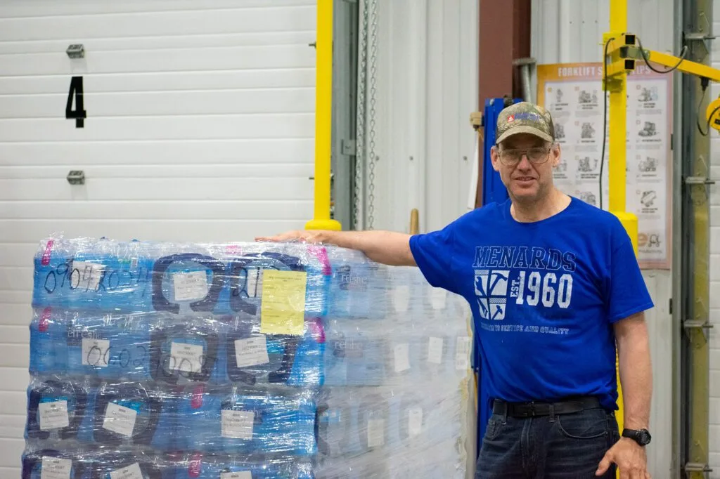 A person stands beside a stacked pallet of bottled water in a warehouse setting.