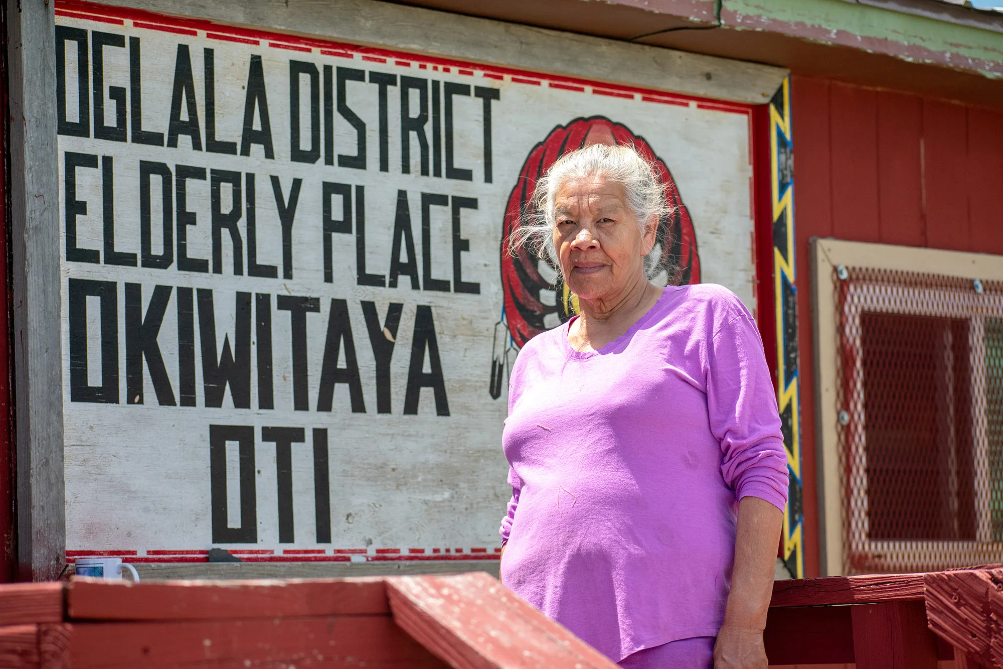 A woman stands in front of a sign that reads “Oglala District Elderly Place Okiwitaya Oti.”