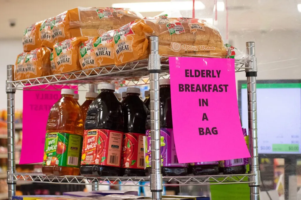 Loaves of honey wheat bread and bottles of juice are displayed with a sign reading "Elderly Breakfast in a Bag."