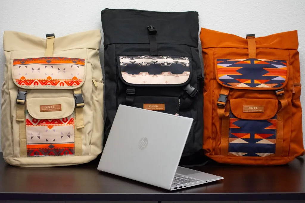 Three patterned backpacks by "KIN CO" and an HP laptop displayed on a table.