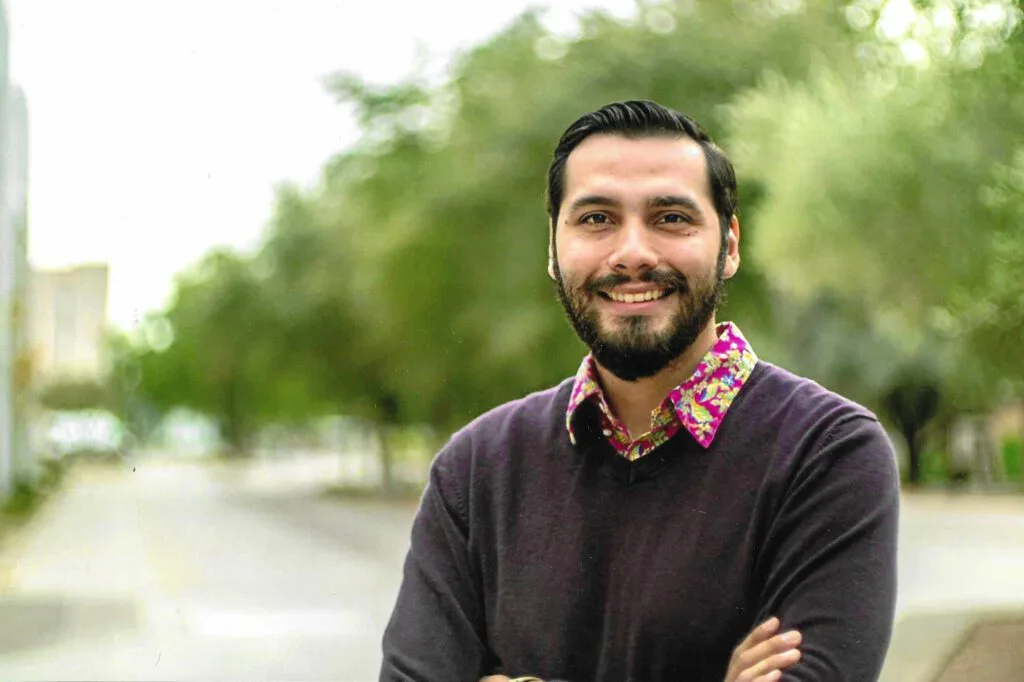 A smiling man with a beard stands outdoors, wearing a dark sweater and a floral-collared shirt with a blurred green background.