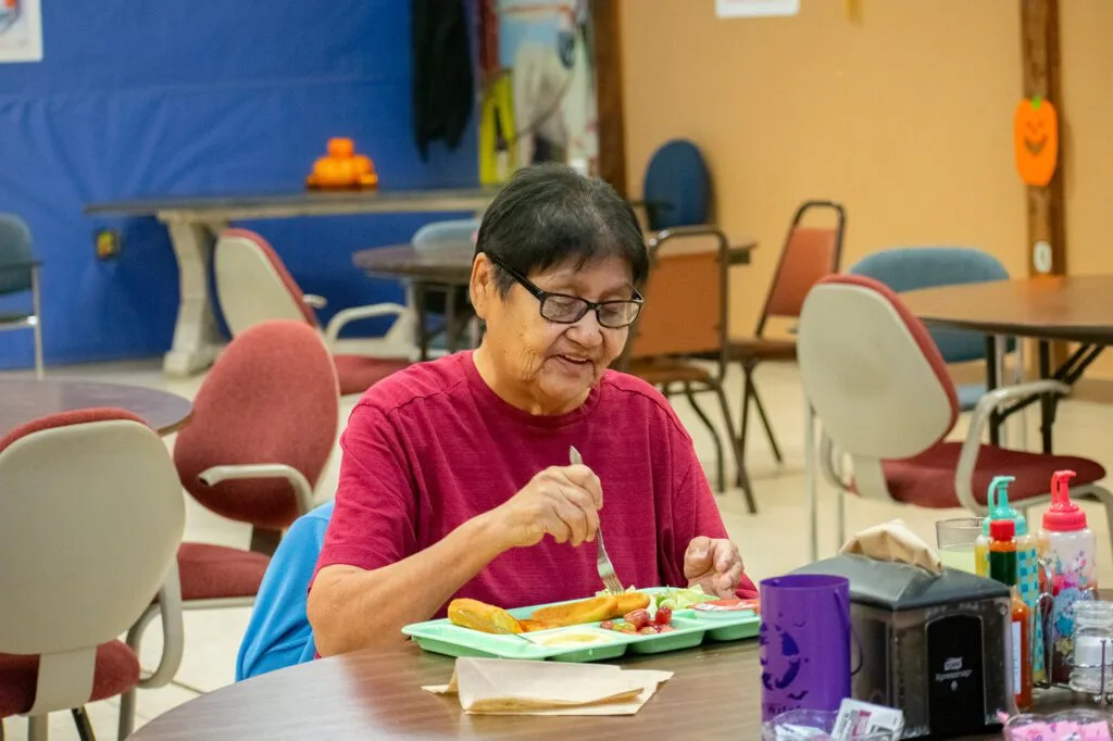 A person is smiling while eating a meal at a table in a communal setting. Northern Plains Reservation Aid is relevant.