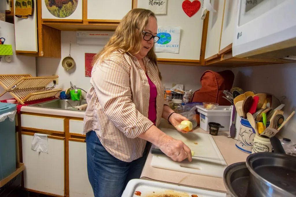 A woman in a kitchen is peeling an onion on a cutting board, surrounded by various kitchen utensils and decor.