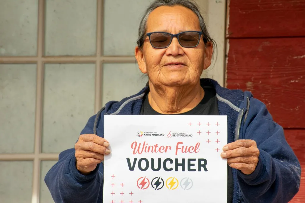 A person holding a "Winter Fuel Voucher" sign from Northern Plains Reservation Aid.