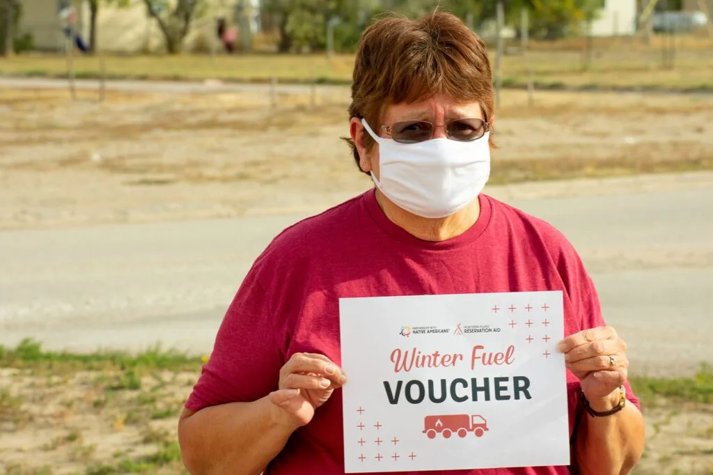 A person wearing a mask holds a "Winter Fuel Voucher" sign with Northern Plains Reservation Aid branding.
