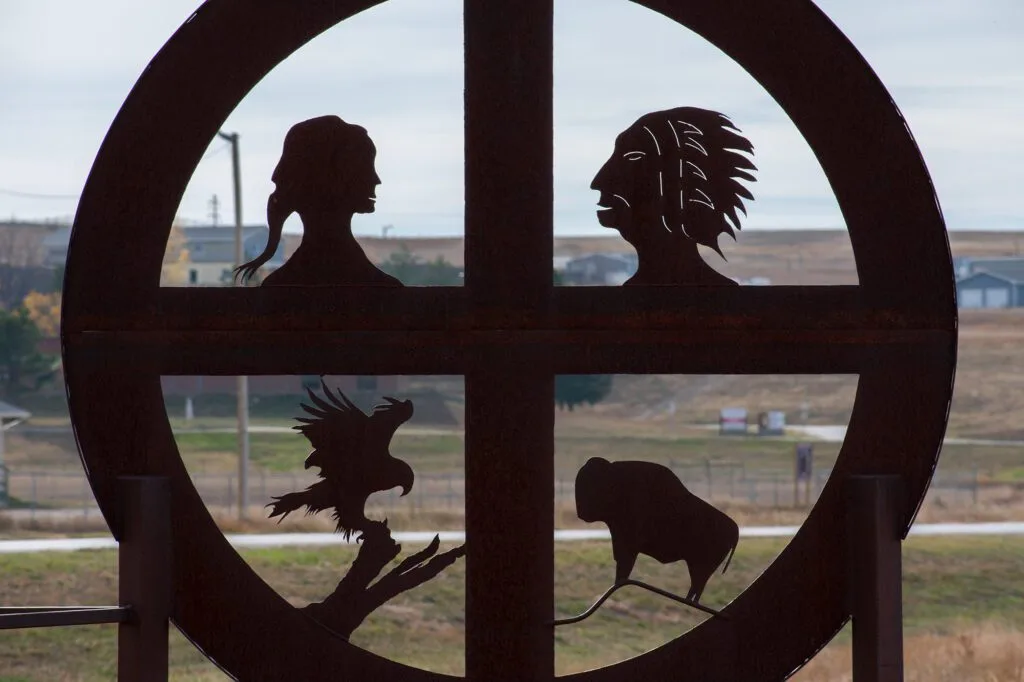 A circular metal sculpture features silhouettes of a woman, a man in a headdress, an eagle, and a bison against a rural backdrop.