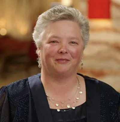 A person in formal attire, wearing earrings and a necklace, stands indoors.
