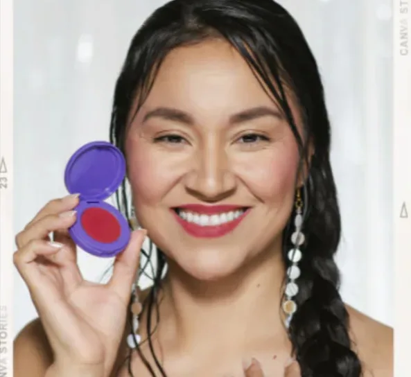A person smiling and holding a round, open container of red makeup.