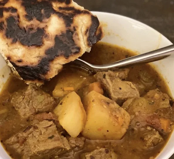 A bowl of hearty stew featuring meat and potatoes, accompanied by a piece of charred flatbread.