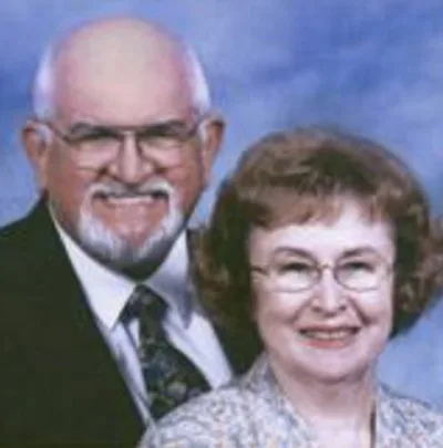 A smiling older couple is dressed in formal attire against a blue background.