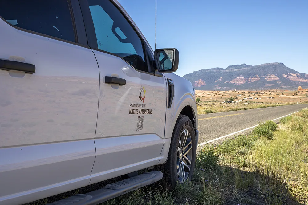 A white truck with the logo "Partnership with Native Americans" parked by a road in a desert landscape with mountains in the distance.