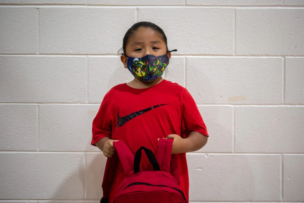 A child wearing a colorful mask and red shirt holds a matching red backpack in front of a white brick wall.