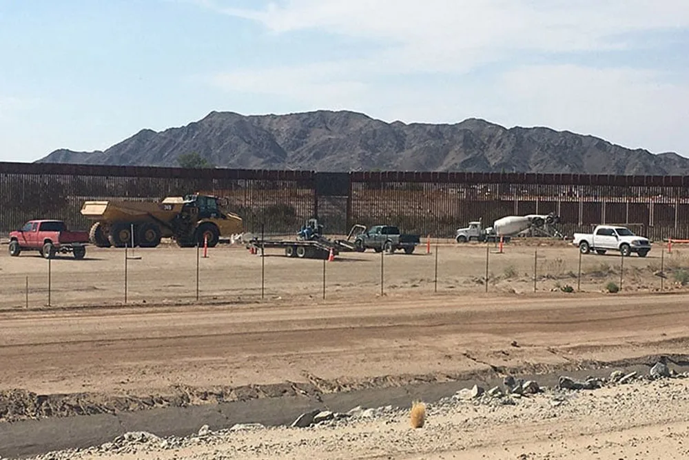 Construction vehicles and trucks near a border fence with mountains in the background.