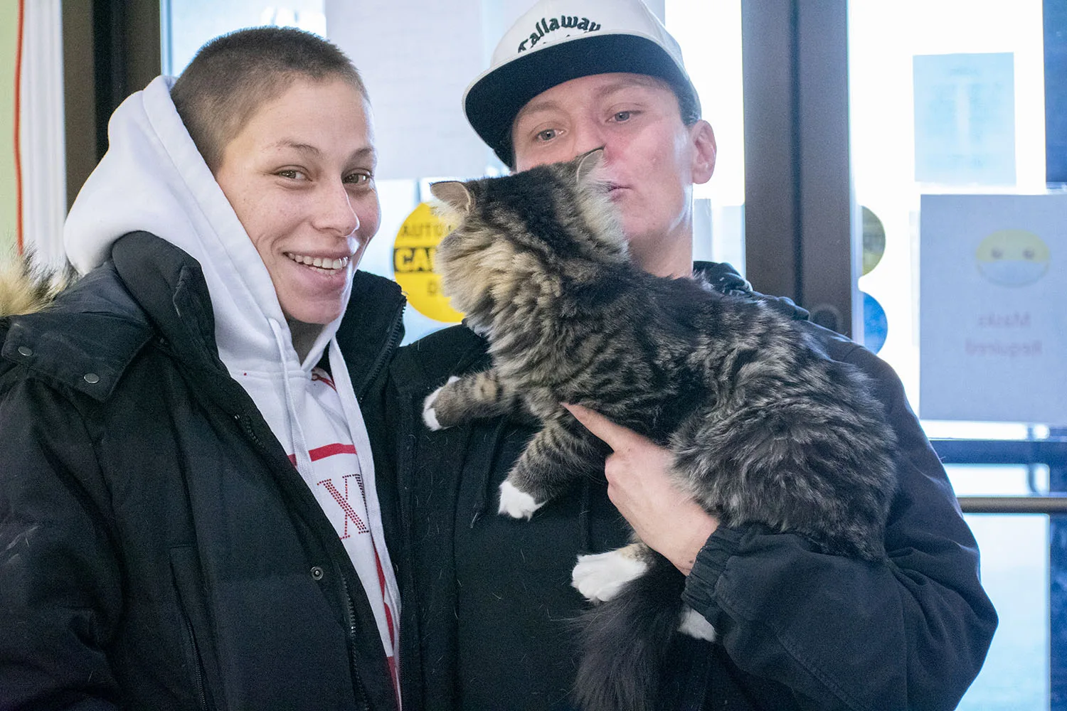 Two people are posing with a fluffy cat indoors, both smiling at the camera.
