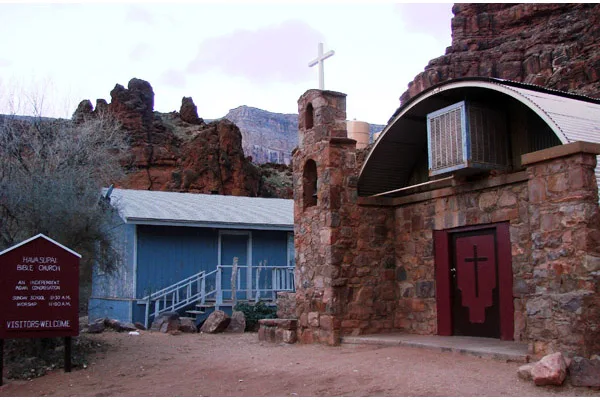 A stone church building with a cross, a blue house, and a sign mentioning "Havasupai Bible Church."