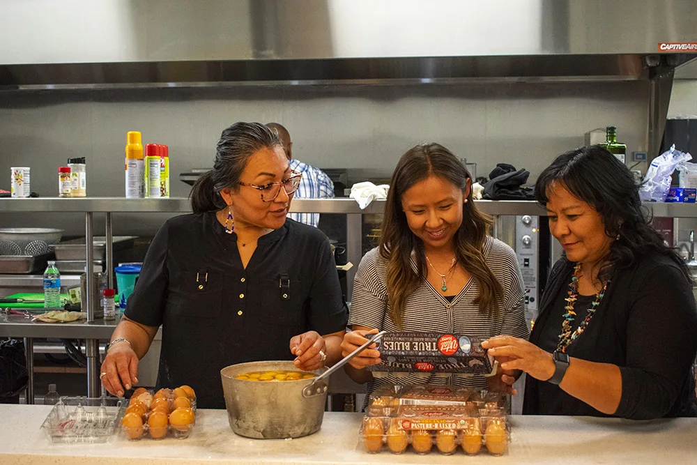 Three women are in a kitchen preparing food, possibly involving eggs. They appear to be organizing cooking materials together.