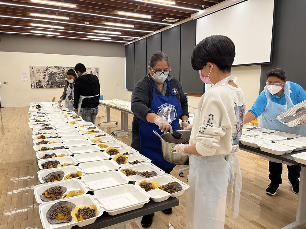 People are preparing meals in a large room with disposable containers on tables.