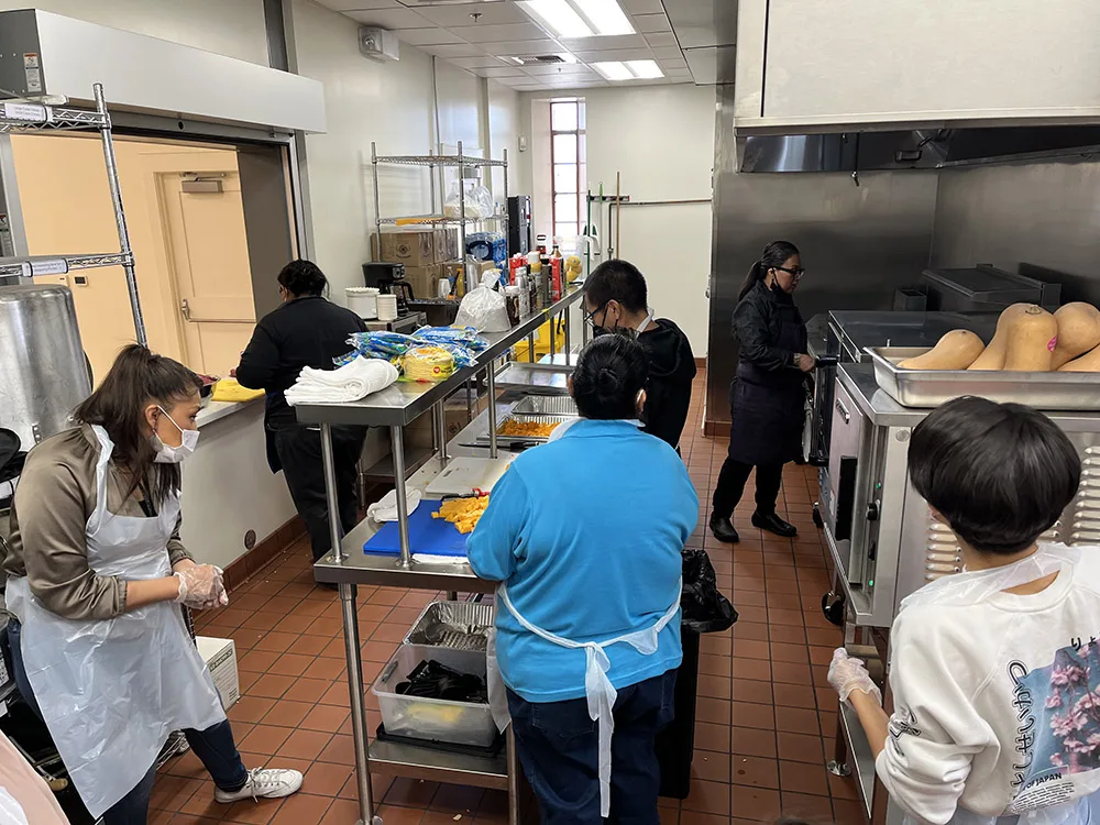 People are preparing food in a commercial kitchen, wearing aprons and masks.