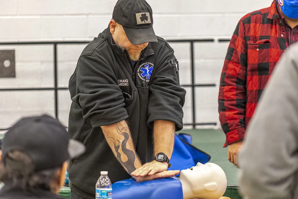 A person in uniform practices CPR on a training dummy, surrounded by others in a classroom or training setting.