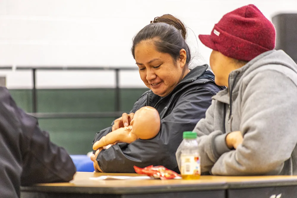 A person holding a baby doll, sitting with two others at a table with snacks and a bottle.