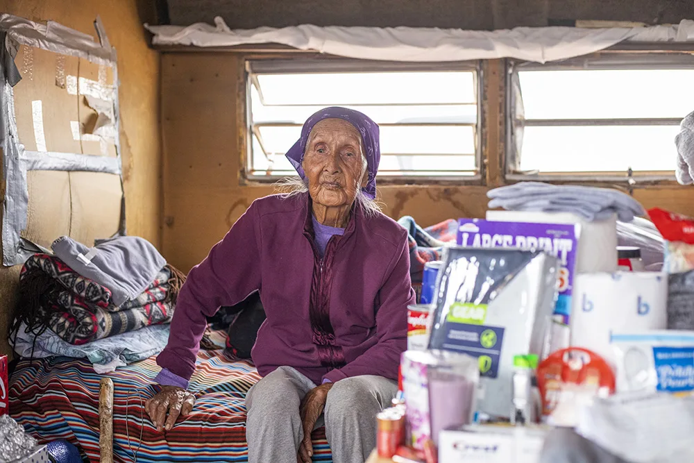 An elderly person sits indoors, surrounded by various supplies and items.