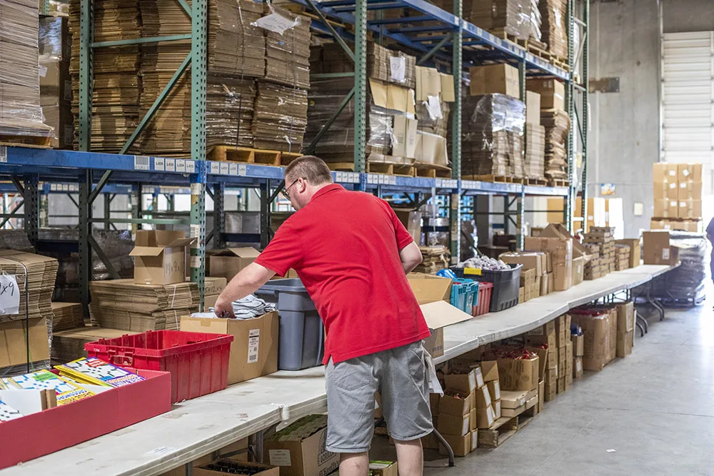 A person in a warehouse setting organizing items on a table surrounded by shelves filled with boxes and supplies.