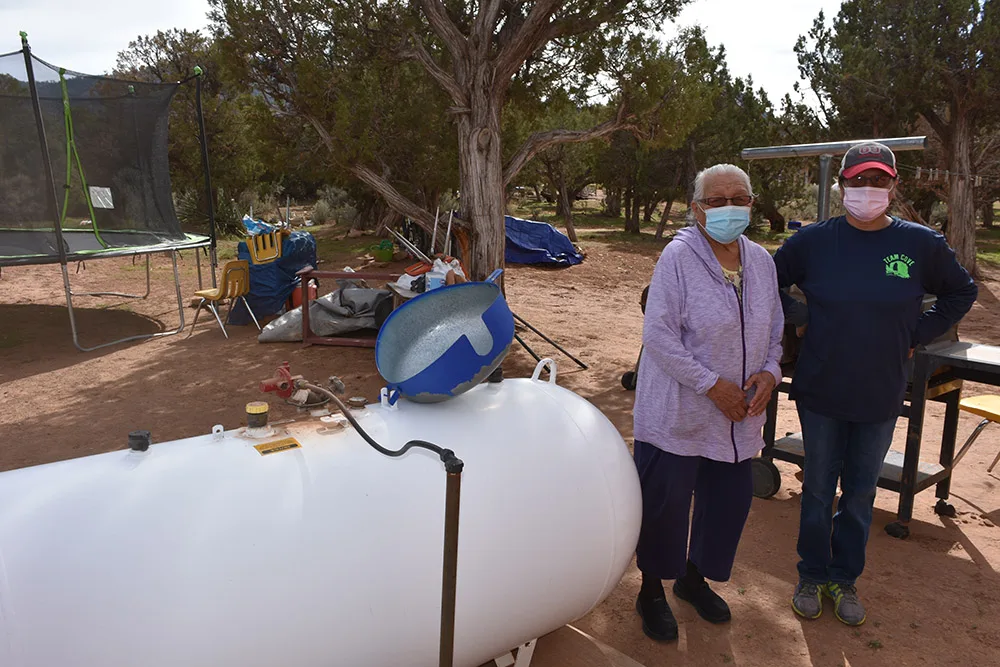 Two people wearing masks stand near a propane tank outdoors, with trees and some recreational equipment in the background.