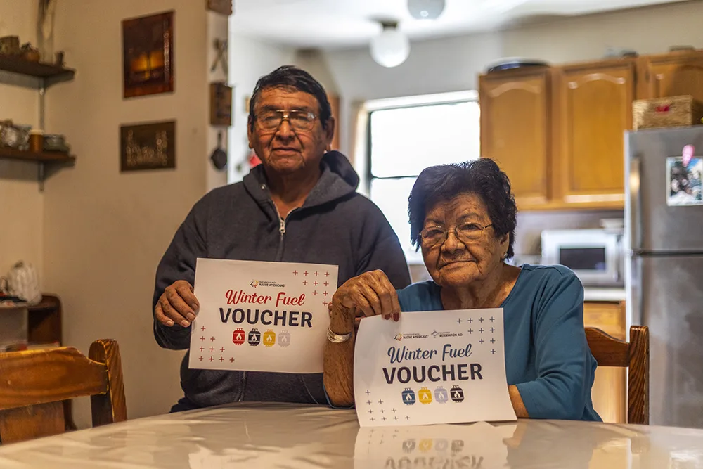 Two people holding "Winter Fuel Voucher" papers at a table in a cozy kitchen setting.