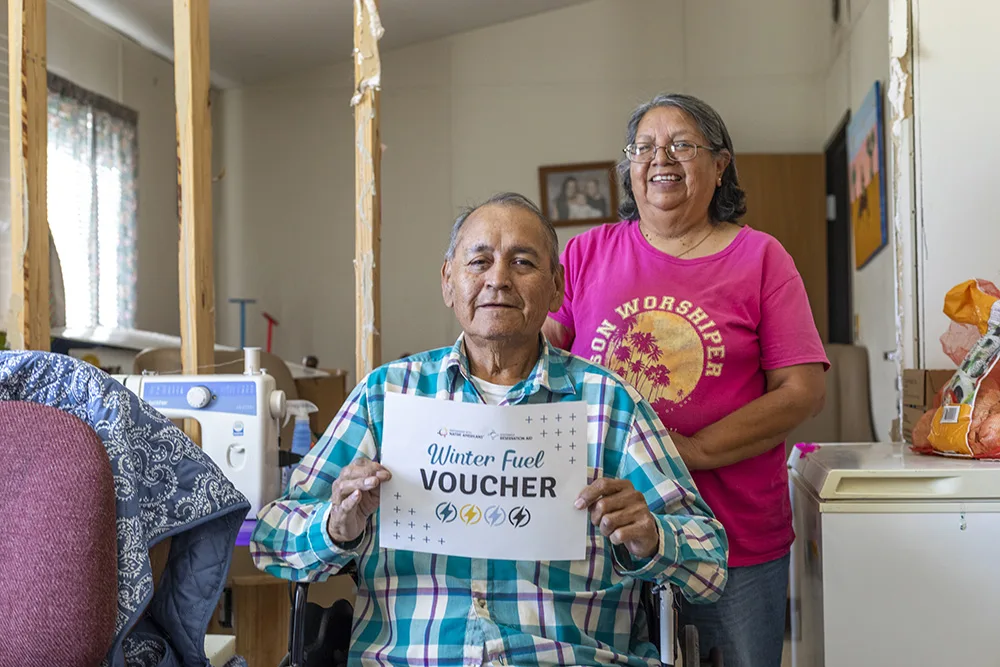 A person is holding a "Winter Fuel Voucher" with another person standing behind them, inside a room.