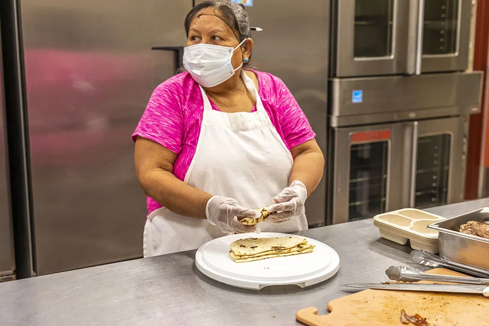 A person in a kitchen is preparing food while wearing a mask and gloves. This could relate to southwest reservation aid.
