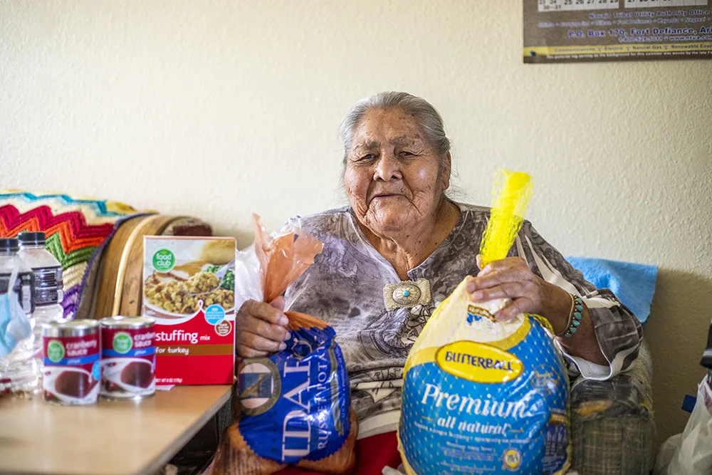An elderly woman is smiling, holding groceries including a turkey, potatoes, stuffing mix, and canned cranberry sauce.