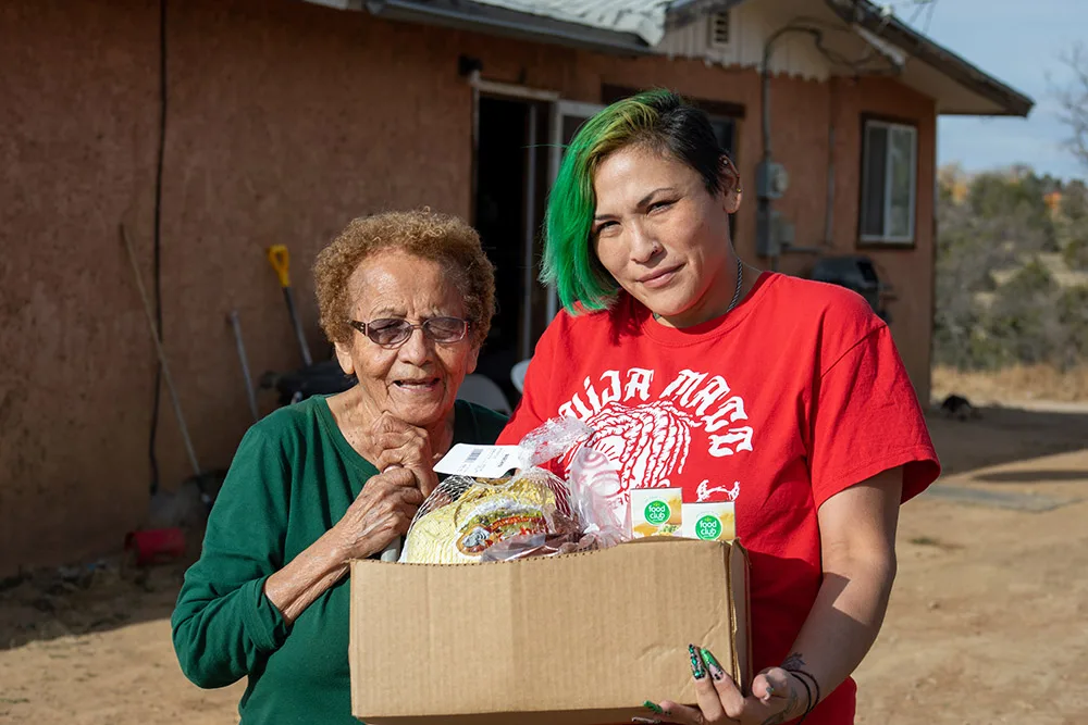 Two people smiling, holding a box of food items outdoors.