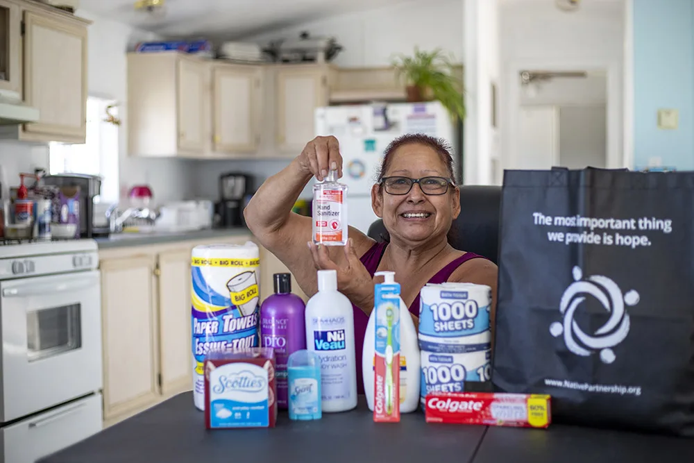 A woman is holding hand sanitizer, surrounded by household items like paper towels and toiletries, alongside a charity bag.