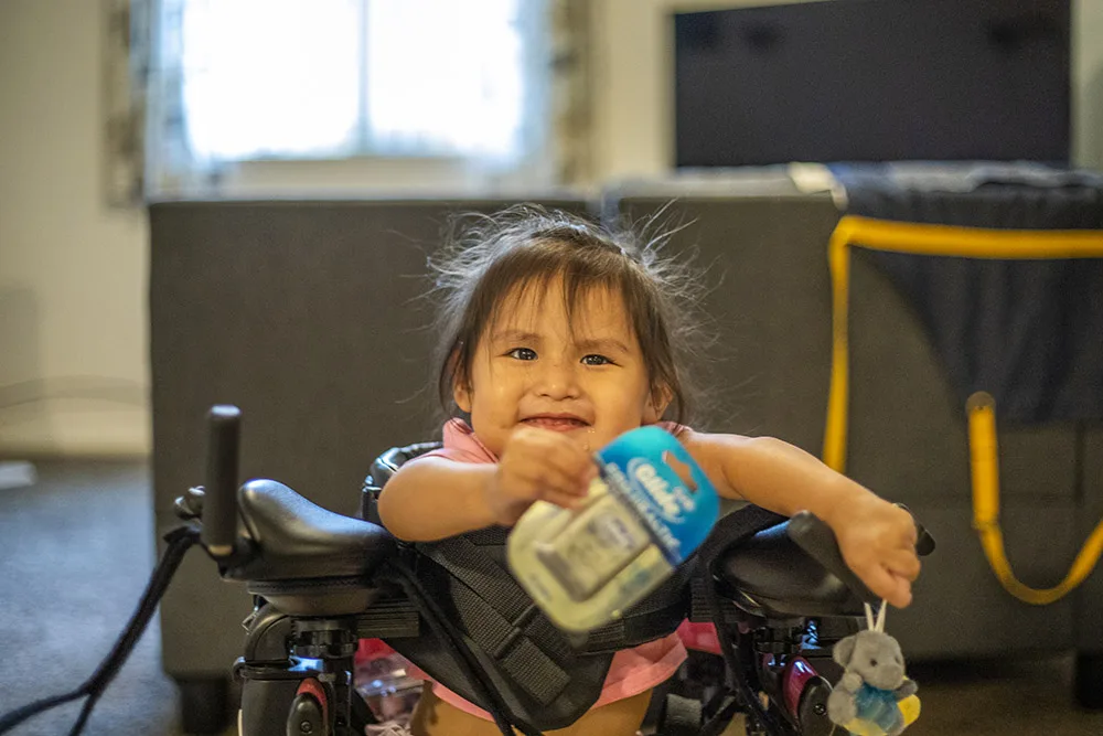 A child with a smile holds a bottle while sitting in a mobility aid indoors.