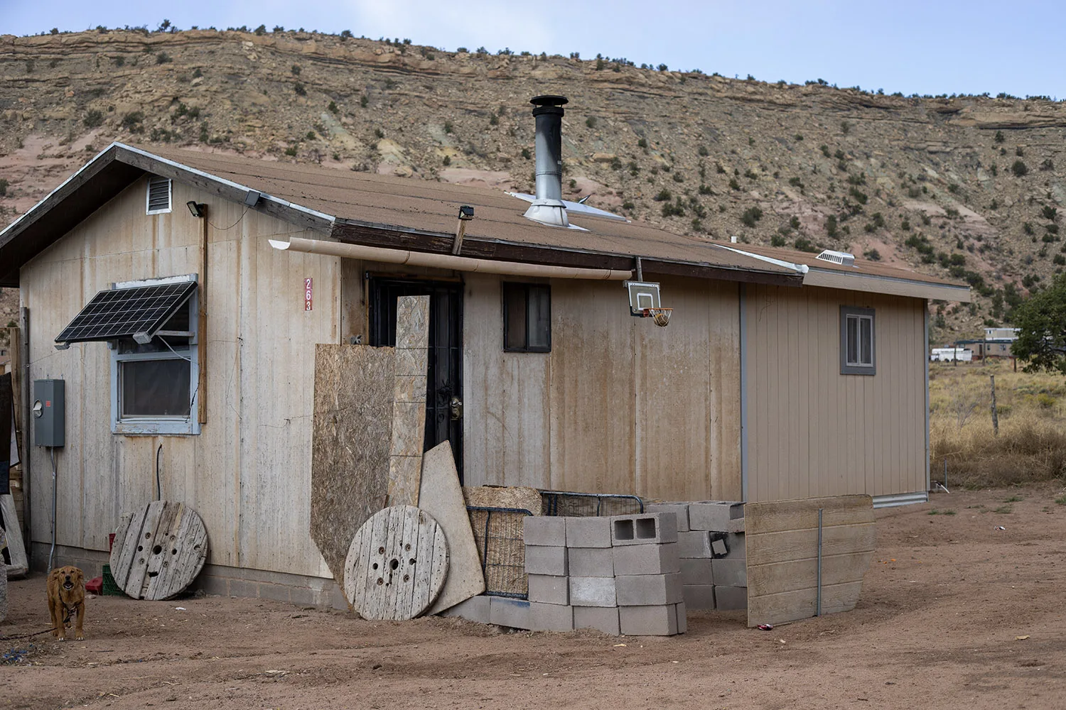 A small, simple house with a solar panel, chimney, cinder blocks, a dog, and desert scenery. Southwest reservation aid is appropriate.