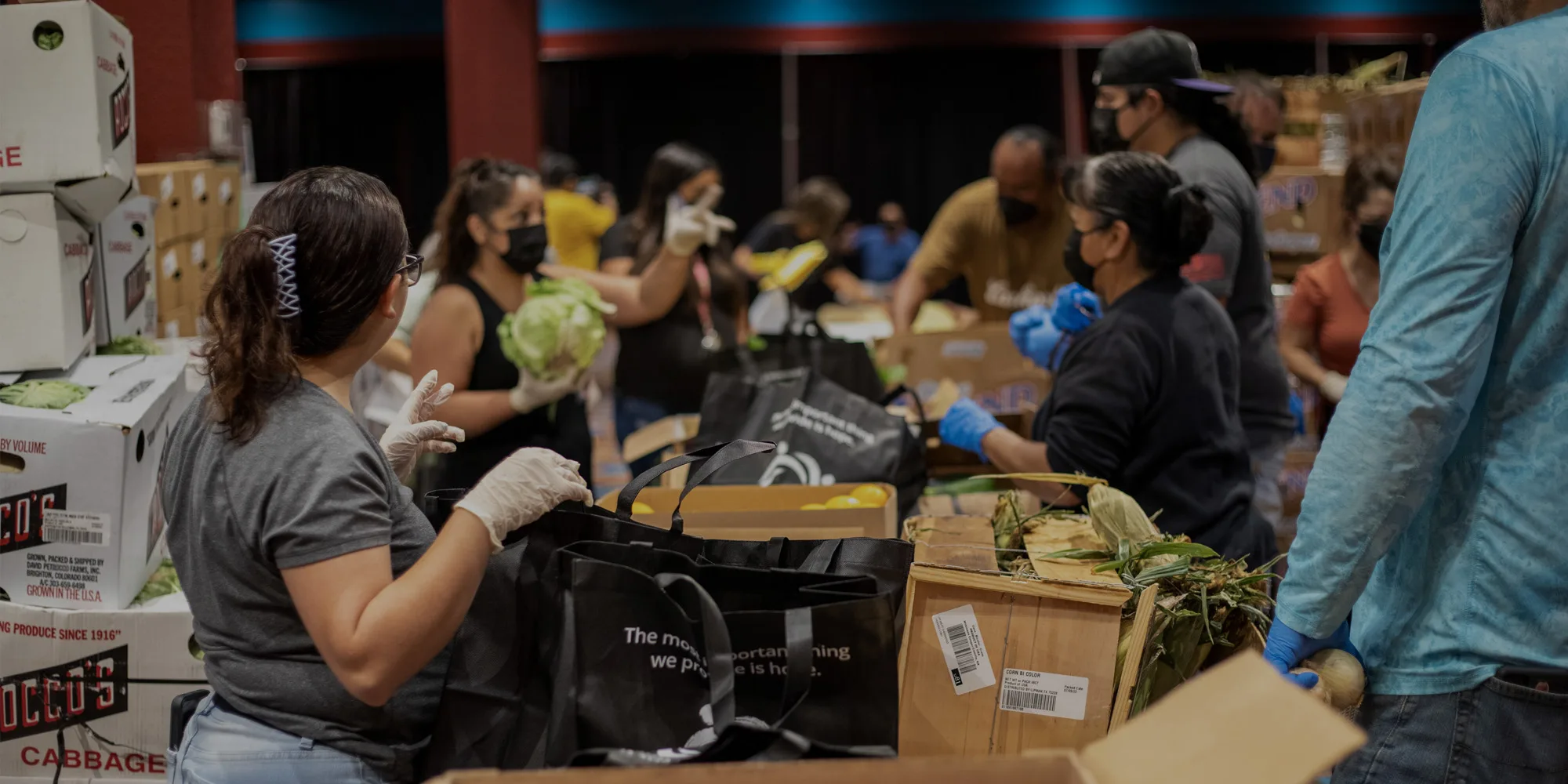 People are gathered, wearing masks and gloves, packing food items into bags.