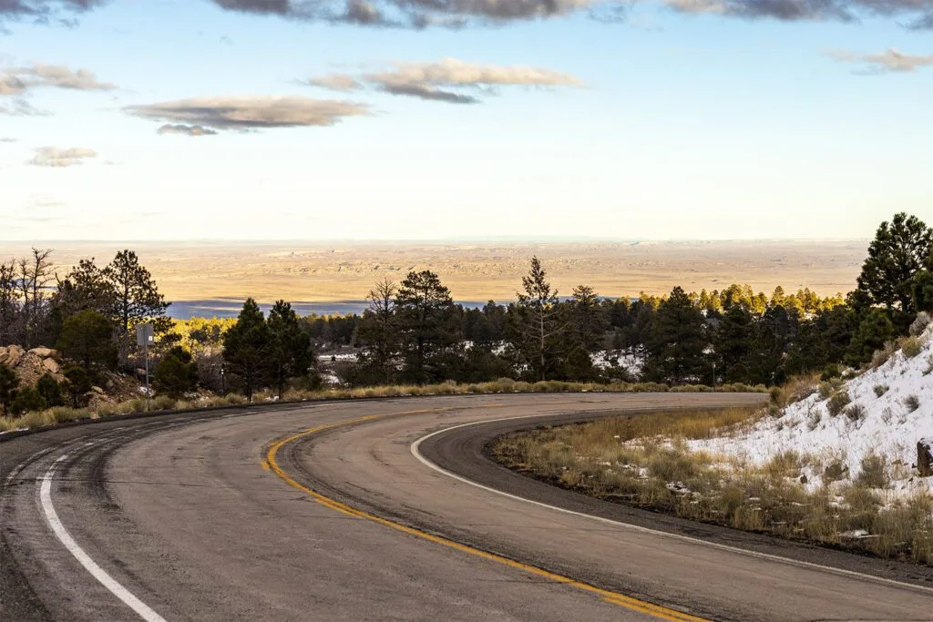 A winding road curves through a forested area with snow patches, leading to a vast, open landscape under a cloudy sky.