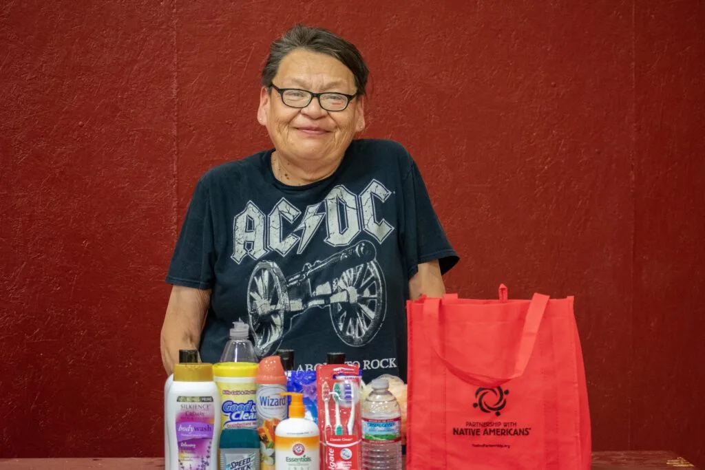 A person stands behind a table displaying personal care items and a red bag from the Partnership with Native Americans.