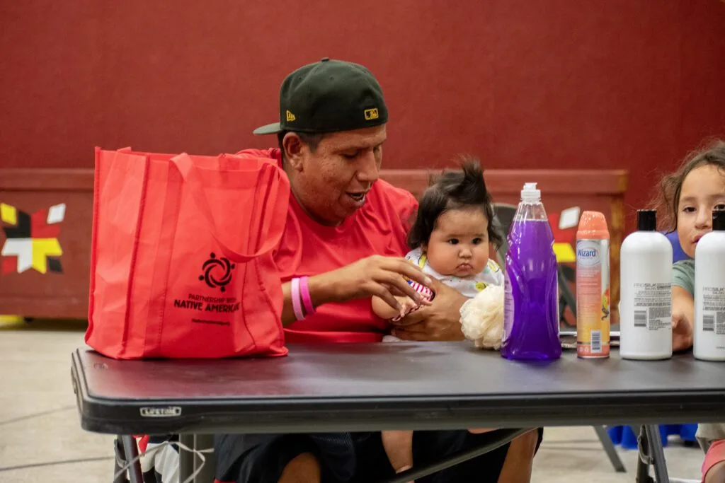 A man and children are seated at a table with household items and a bag labeled "Partnership With Native Americans."