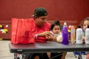 A man and children are seated at a table with household items and a bag labeled "Partnership With Native Americans."