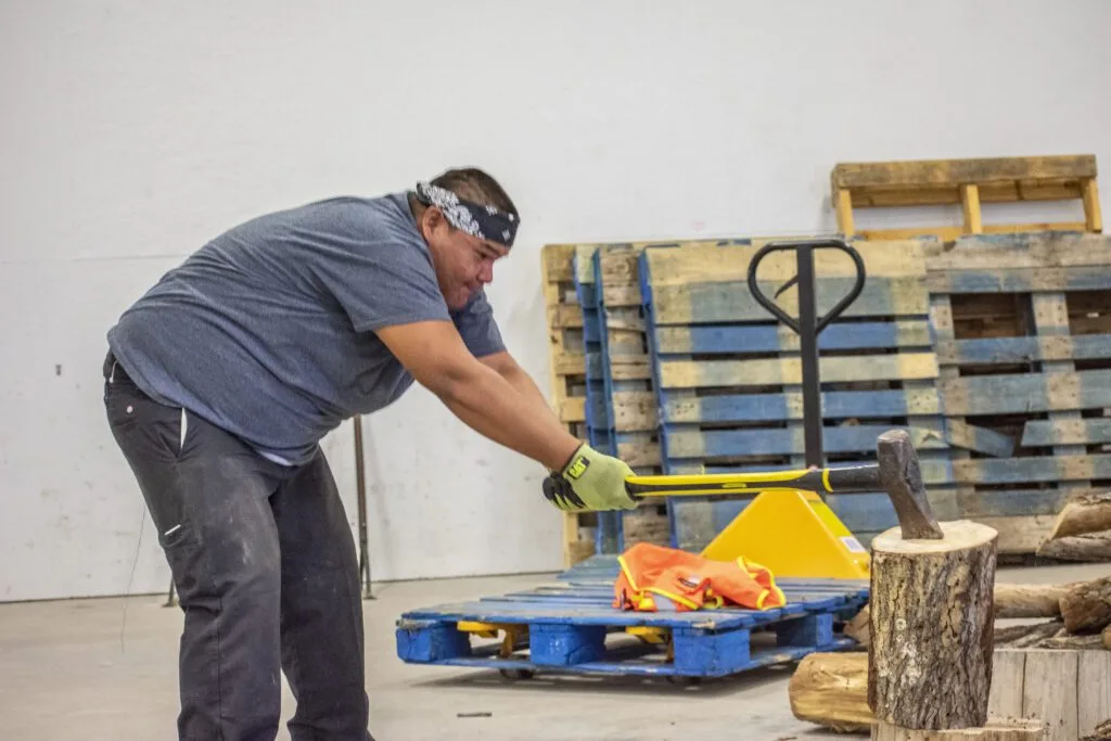 A person is chopping wood indoors with an axe, surrounded by pallets and logs.