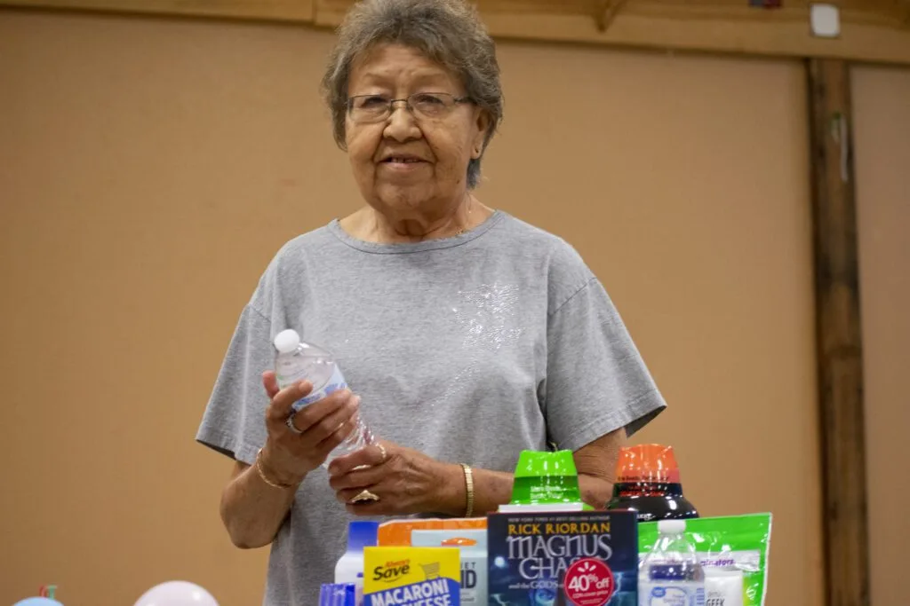 An elderly person is holding a water bottle near a table with groceries and household items.