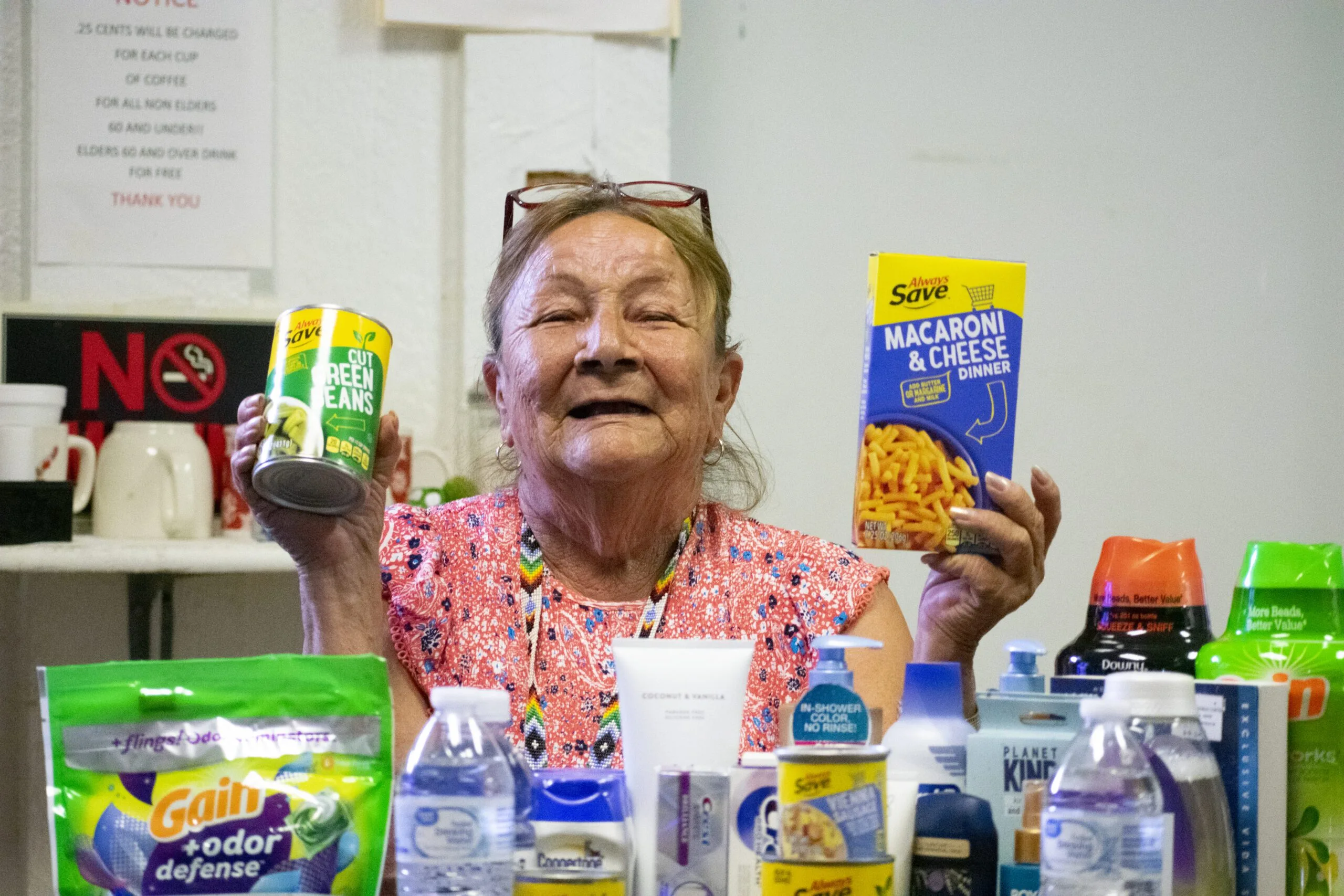 An elderly woman holds a can of green beans and a box of macaroni and cheese, surrounded by various groceries.
